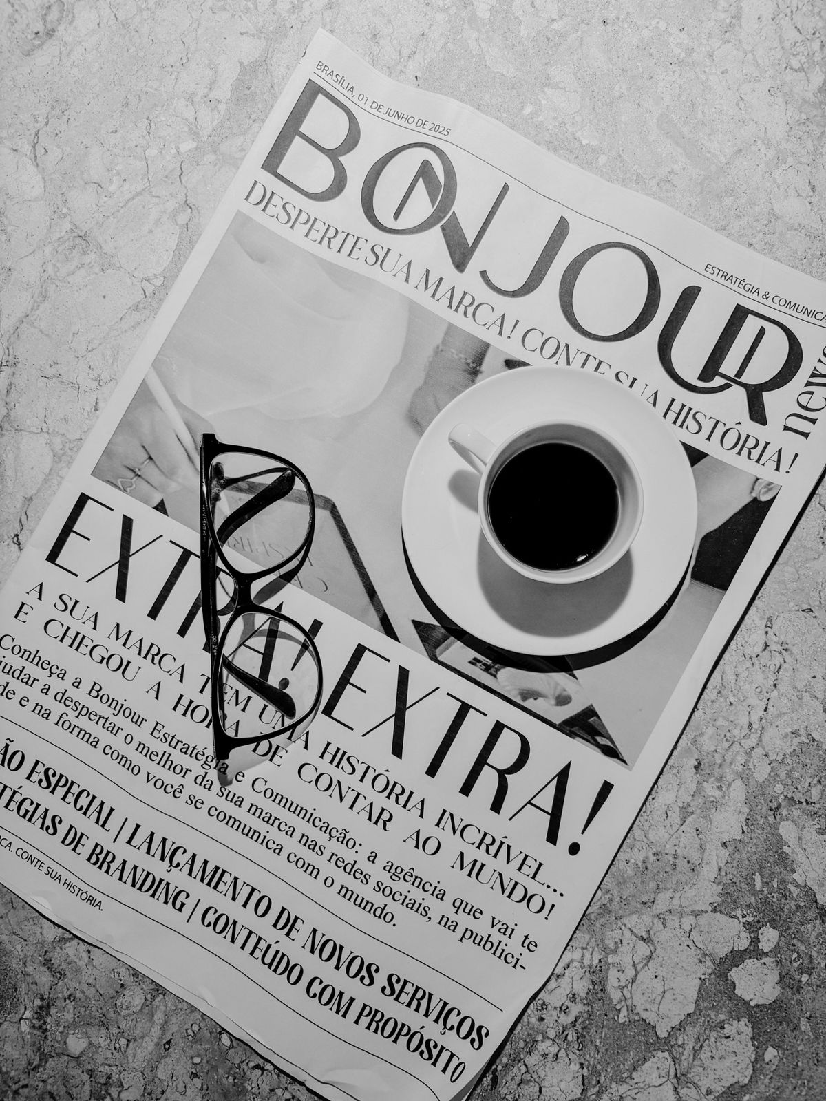 Jornal com a marca Bonjour, xícara de café e óculos sobre a mesa, em preto e branco